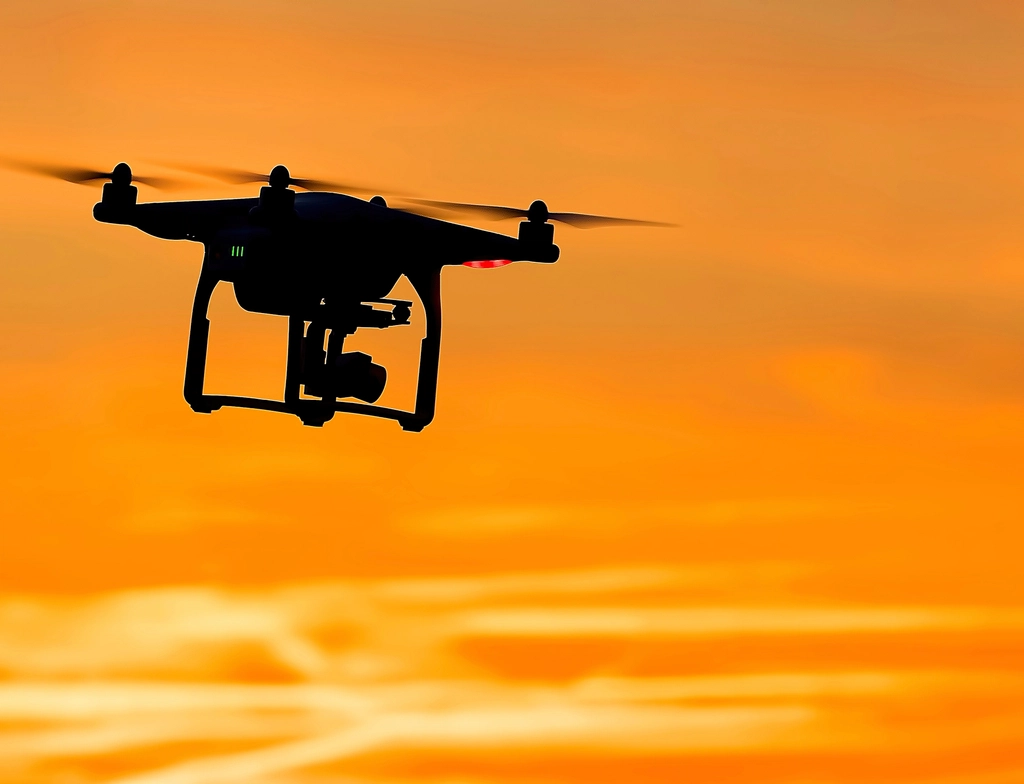 Silhouette of a camera drone flying against an orange sunset sky
