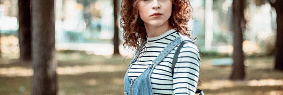 A young woman with curly hair standing in a park in a striped shirt and overalls