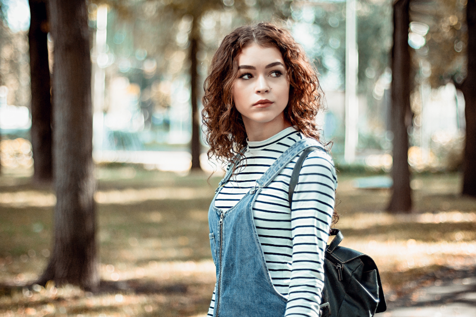 A young woman with curly hair standing in a park in a striped shirt and overalls