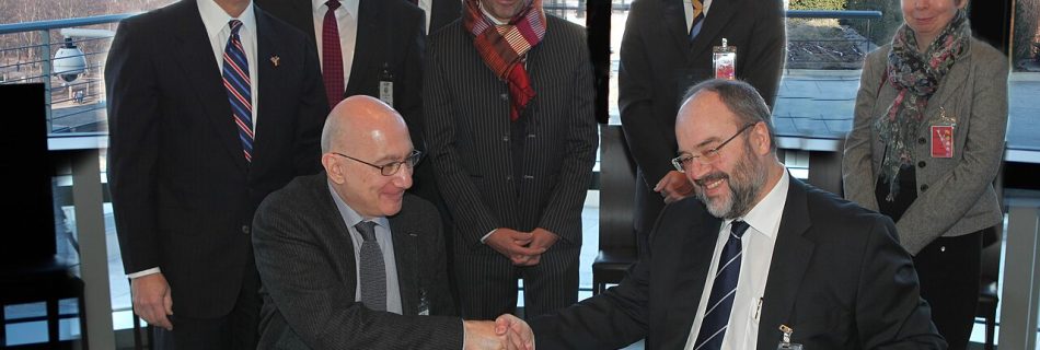 Two men shaking hands over signed agreement documents while colleagues stand behind them