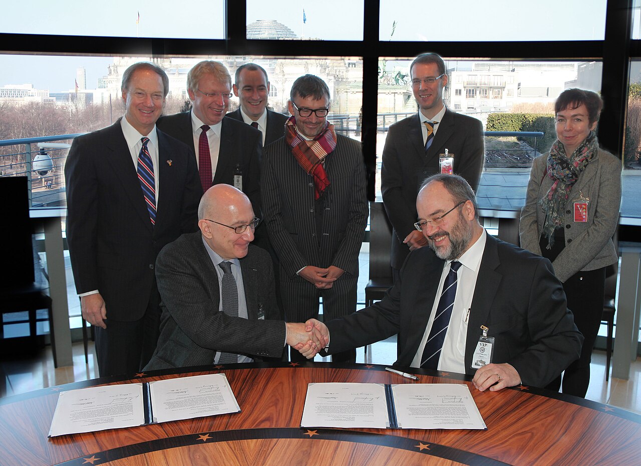 Two men shaking hands over signed agreement documents while colleagues stand behind them